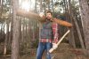 Ein Mann im Wald, der ein Holzstamm auf der rechten Schulter trägt und in der linken Hand ein Beil hält.