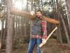 Ein Mann im Wald, der ein Holzstamm auf der rechten Schulter trägt und in der linken Hand ein Beil hält.
