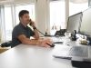 Ein Mann sitzt vor 2 Computerbildschirmen und hat den Telefonhörer in der linken Hand.