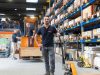 Ein Mann steht mit einem Paletthubwagen in einem grossen Logistiklager. Im Hintergrund stehen zwei Männer und unterhalten sich. Einer von den Männern steht auf einem Gabelstapler.